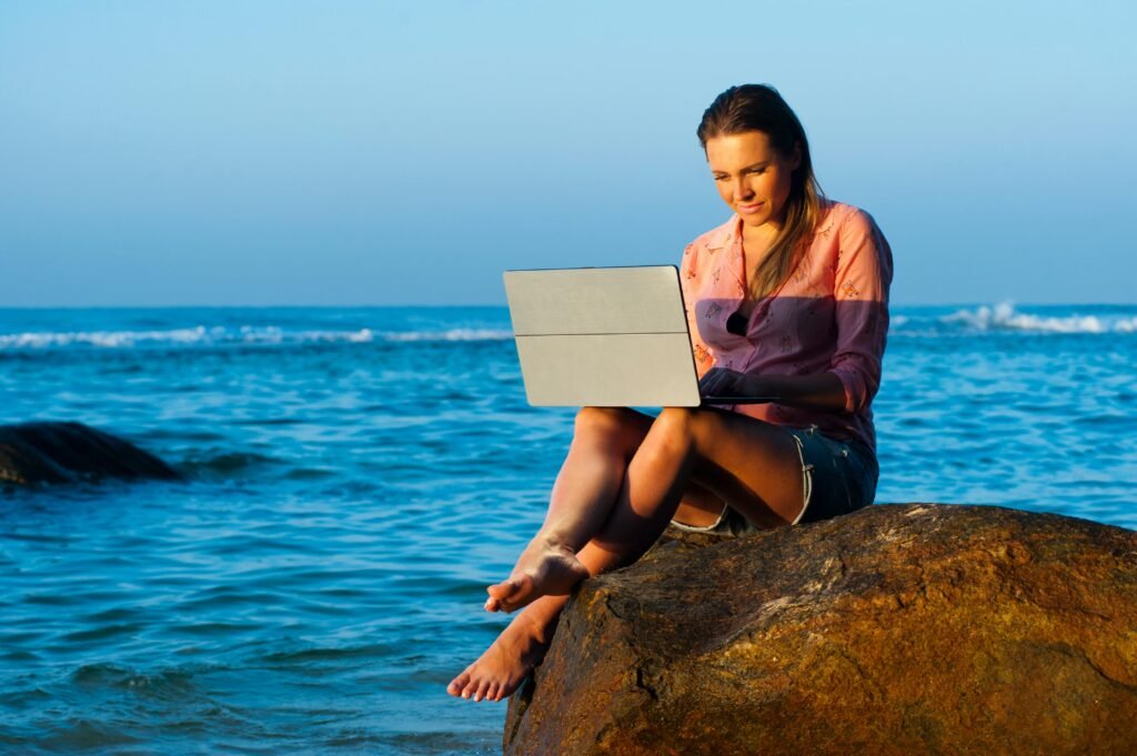 A woman relaxes on a rock by the sea in Sri Lanka, using her laptop during a summer day.
